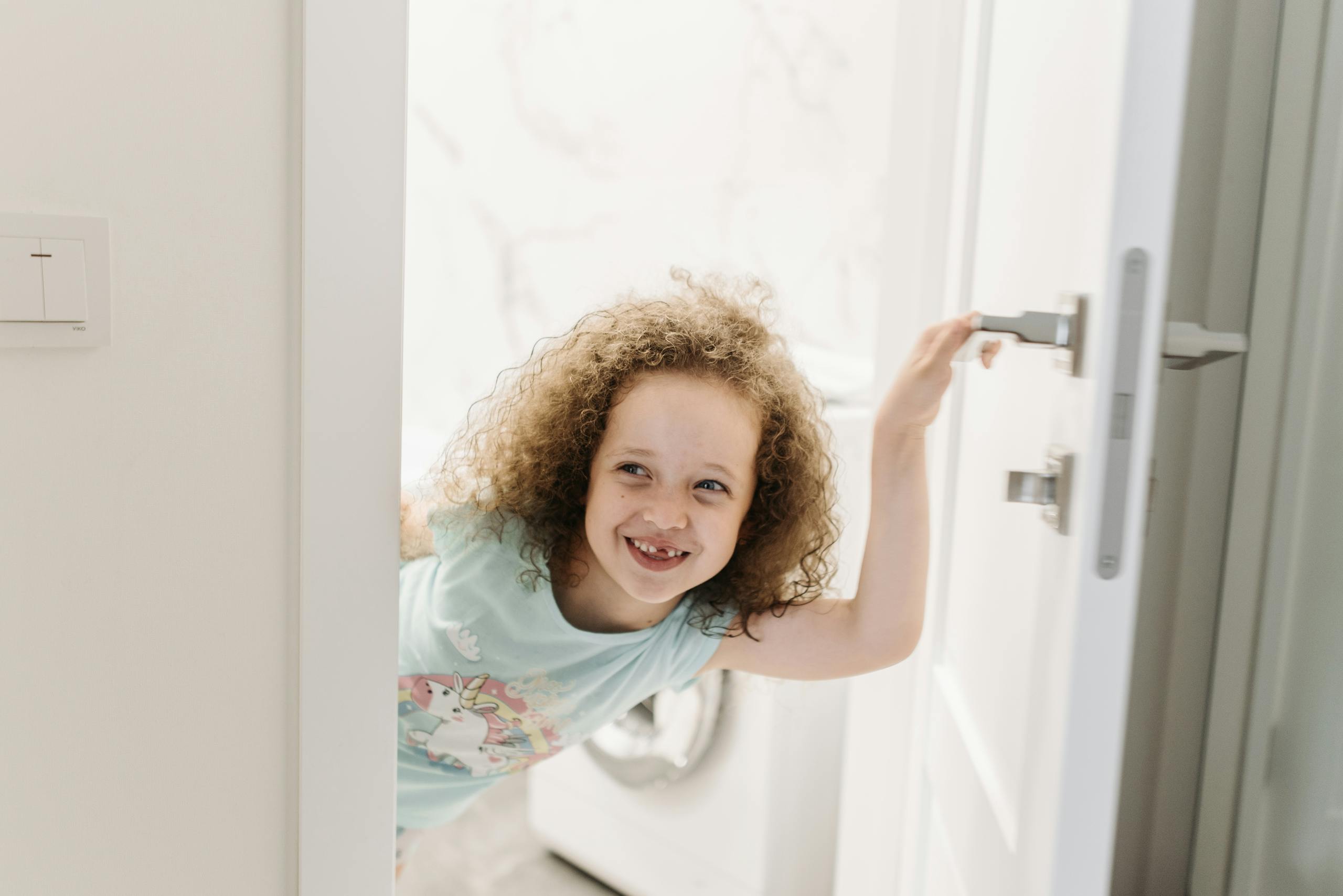 Joyful child with curly hair smiling as she peeks through a doorway indoors.